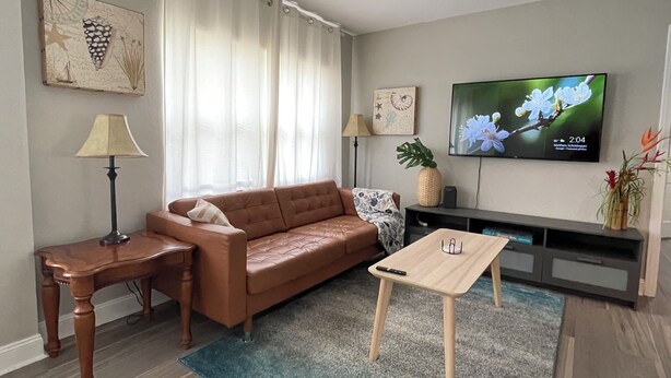 Un salón con un sofá de cuero marrón, una mesa de centro de madera, un televisor en la pared y una mesita auxiliar con una lámpara.