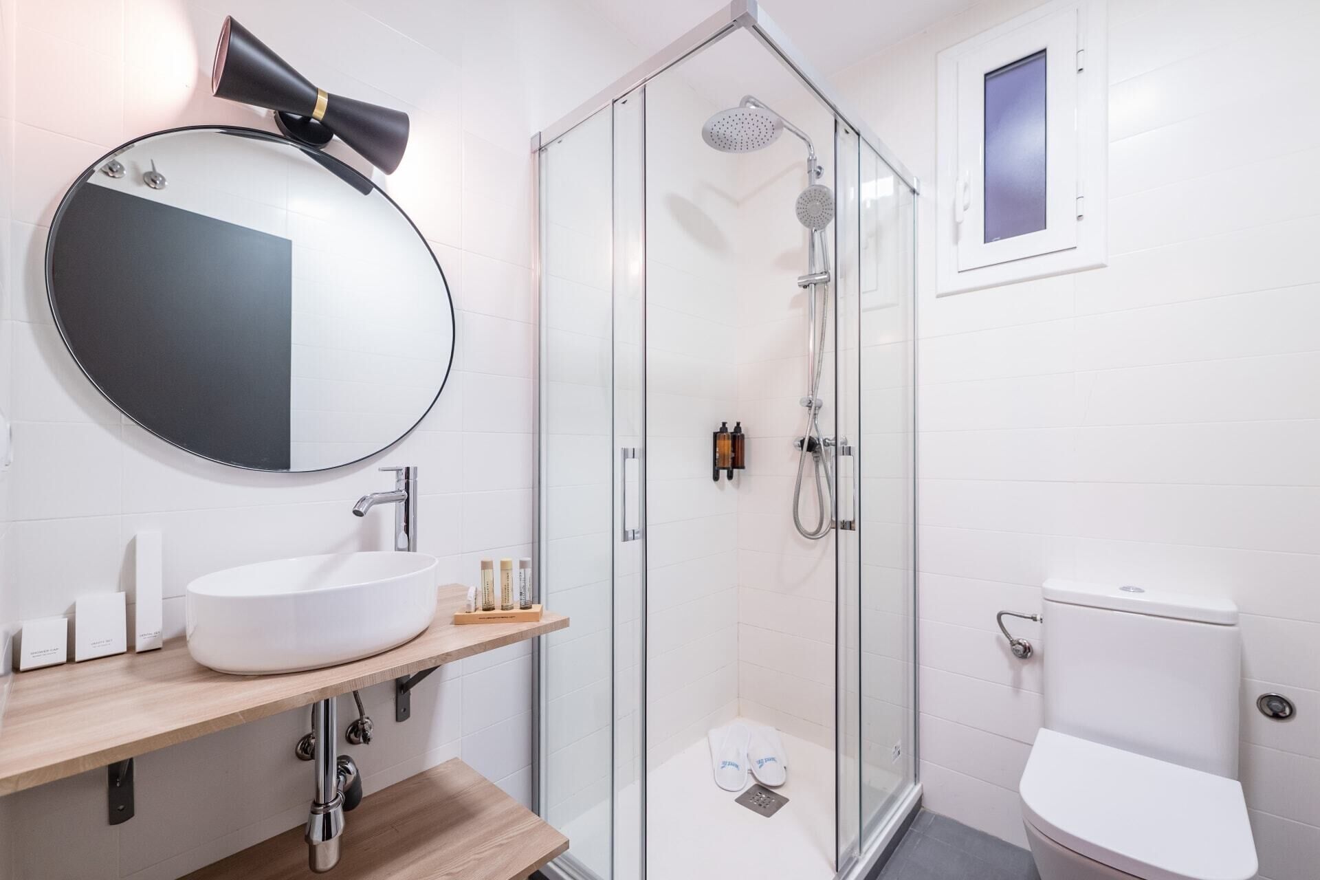 A modern bathroom with a round mirror, a white sink, a glass-enclosed shower, and a toilet.