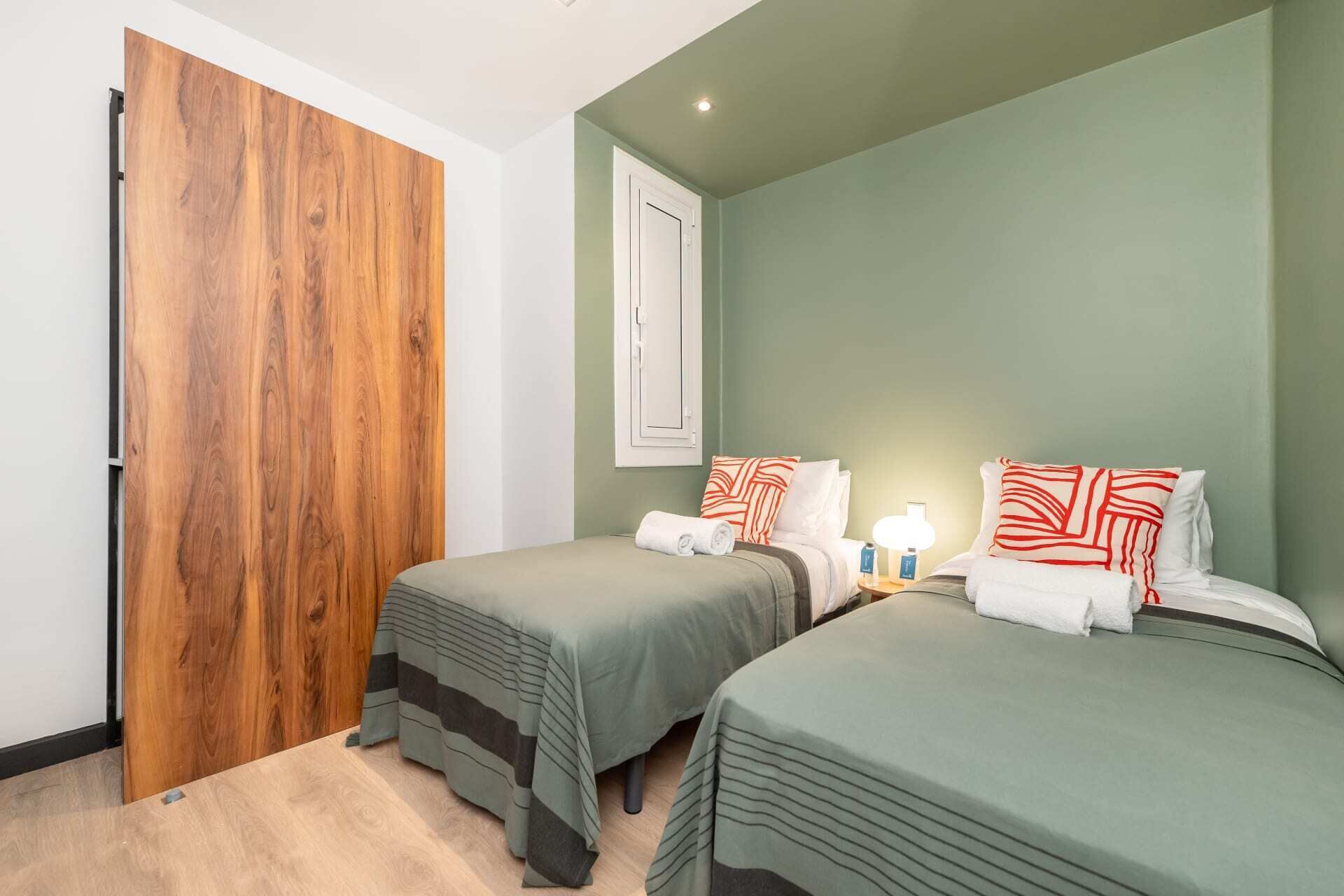 Une chambre d’hôtel avec deux lits simples, une porte en bois, une fenêtre et une table de chevet avec une lampe.