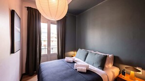 A modern bedroom with a large bed, dark walls, a hanging light fixture, and a view of buildings through the window.