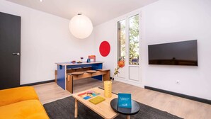 A modern living room with a sofa, a coffee table, a TV, a dining area with a table and chairs, and a large window.