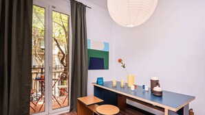 Una habitación moderna con un escritorio de madera, dos taburetes, un ventanal con cortinas, una lámpara colgante y vistas a un balcón con mobiliario exterior.