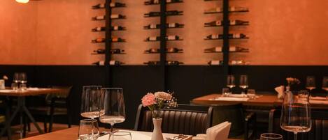 A restaurant with tables set for dining, a wall of wine bottles, and a vase with flowers on the table.