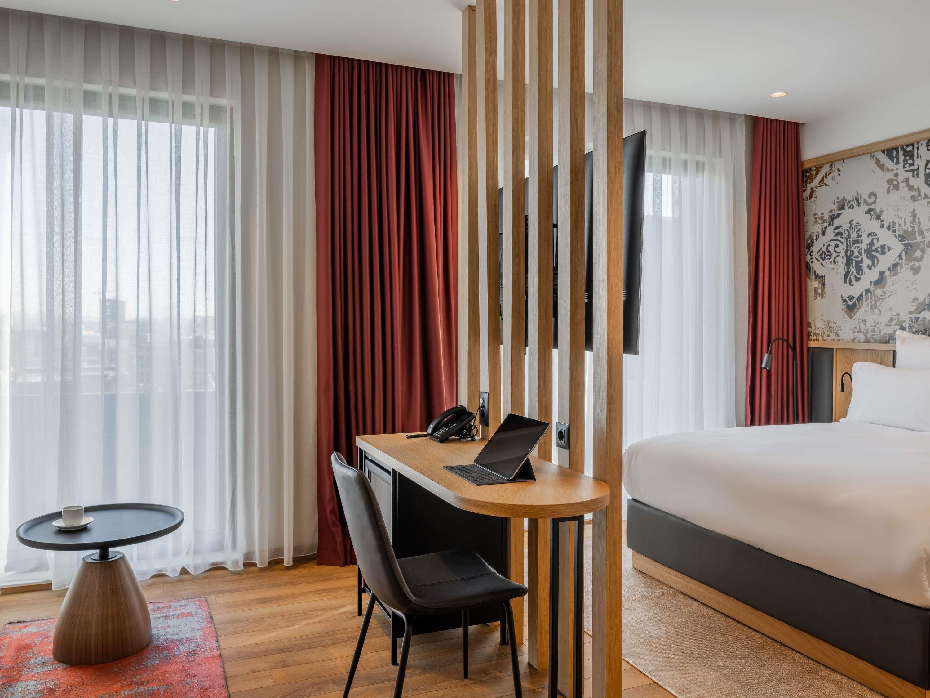 A hotel room with a bed, a desk with a laptop, a chair, a phone, and a TV.