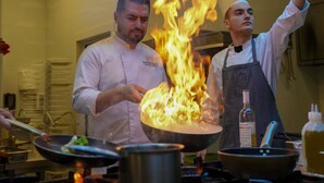 Köche führen eine Flambierung in einer Küche mit Edelstahlgeräten und einer gefliesten Spritzschutzwand durch.