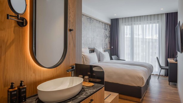 Ein modernes Hotelzimmer mit einem großen, ovalen Spiegel, einem runden, weißen Waschbecken, einem Bett mit weißer Bettwäsche, einem Schreibtisch mit Stuhl und einem großen Fenster mit transparenten Vorhängen.