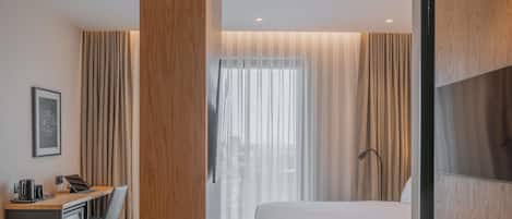 A modern hotel room with a wooden desk, a bed, and a large window with sheer curtains.