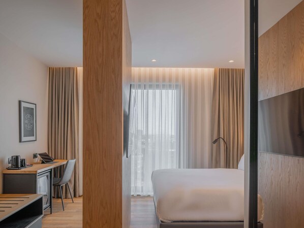A modern hotel room with a wooden desk, a bed, and a large window with sheer curtains.