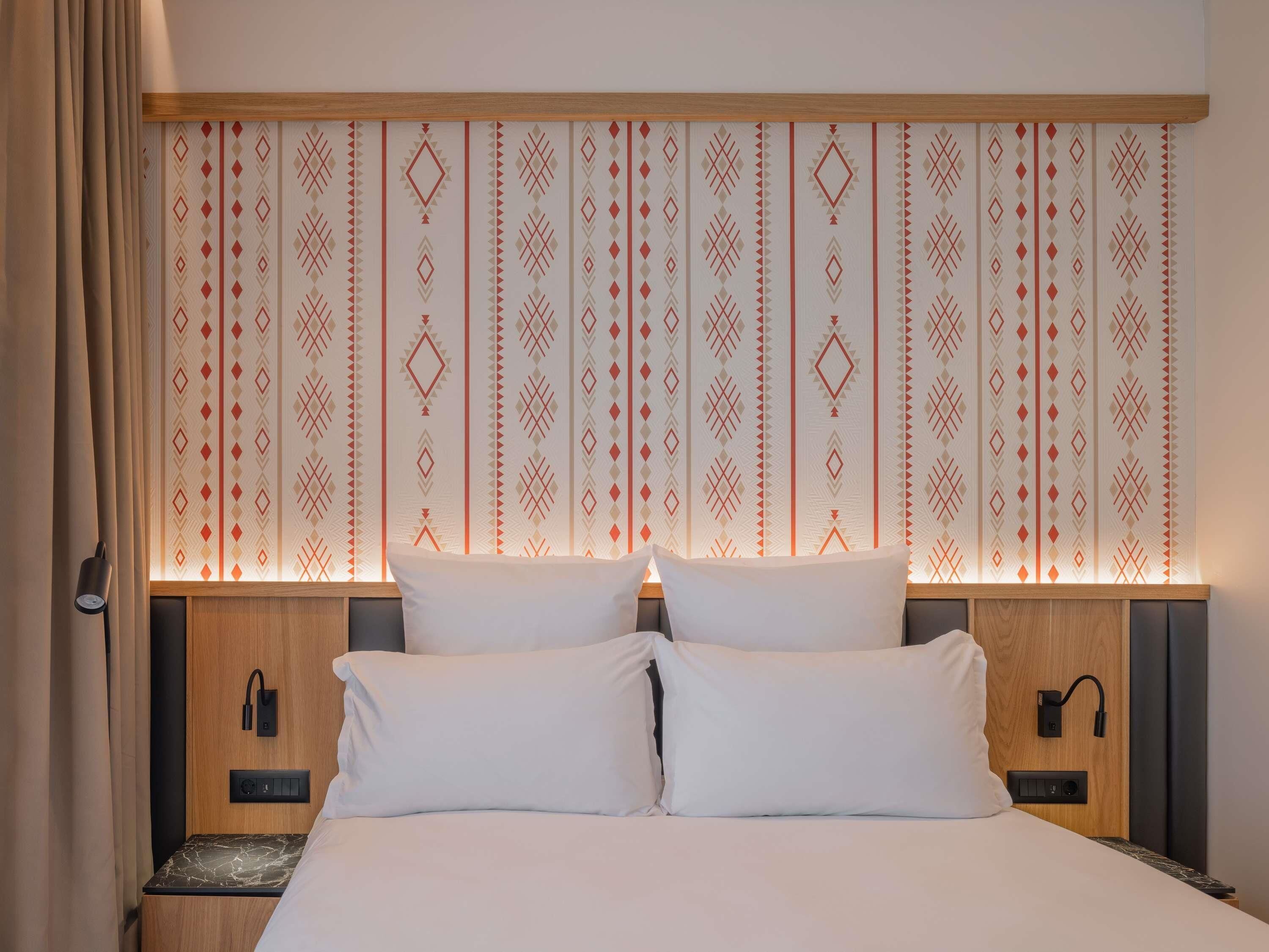 Una habitació d'hotel amb llit, dues taules de nit, una tauleta auxiliar, una làmpada de paret i un capçal de llit amb disseny.