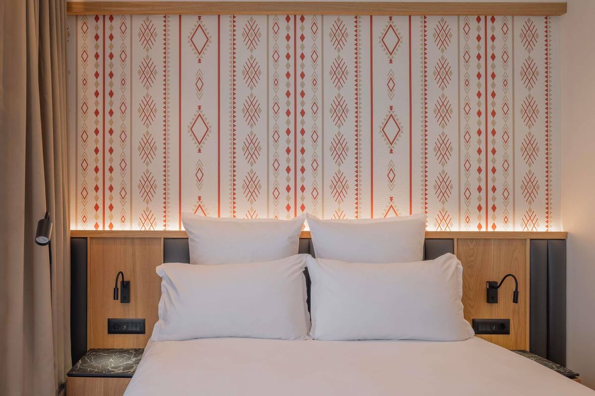 Une chambre d’hôtel comprenant un lit, deux tables de chevet, une petite table, une lampe fixée au mur et une tête de lit à motifs.