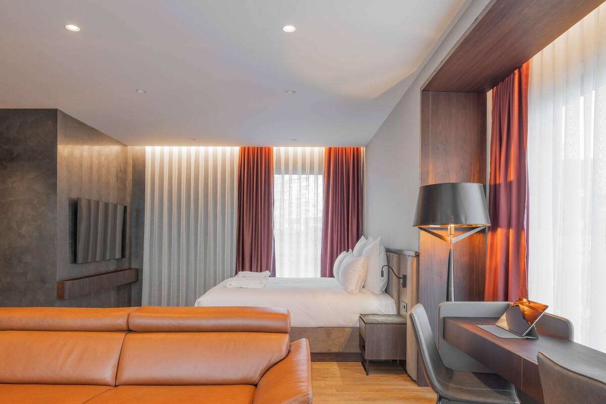 Une chambre d’hôtel moderne équipée d’un lit, d’un canapé, d’un bureau avec un ordinateur portable, d’une lampe et d’un téléviseur fixé au mur.