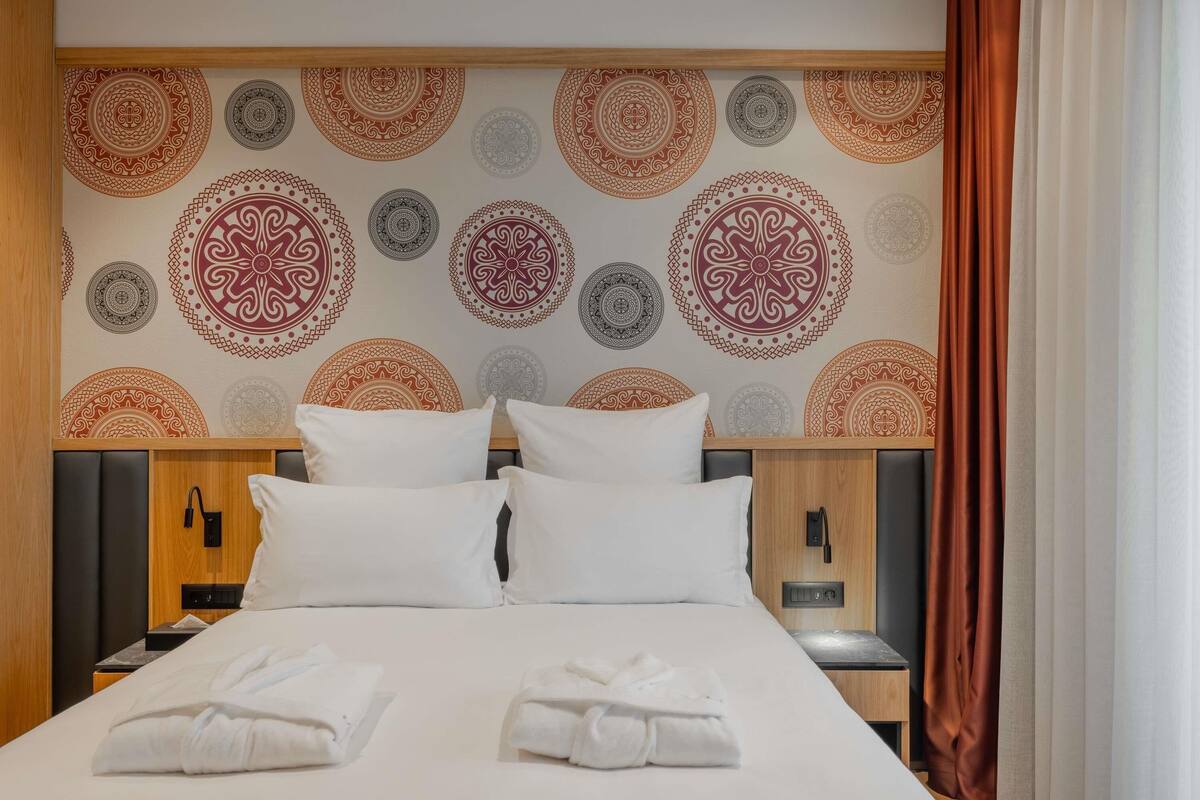 Une chambre d’hôtel comprenant un lit, des oreillers, une table de chevet, une tête de lit en bois et un mur orné de motifs circulaires.