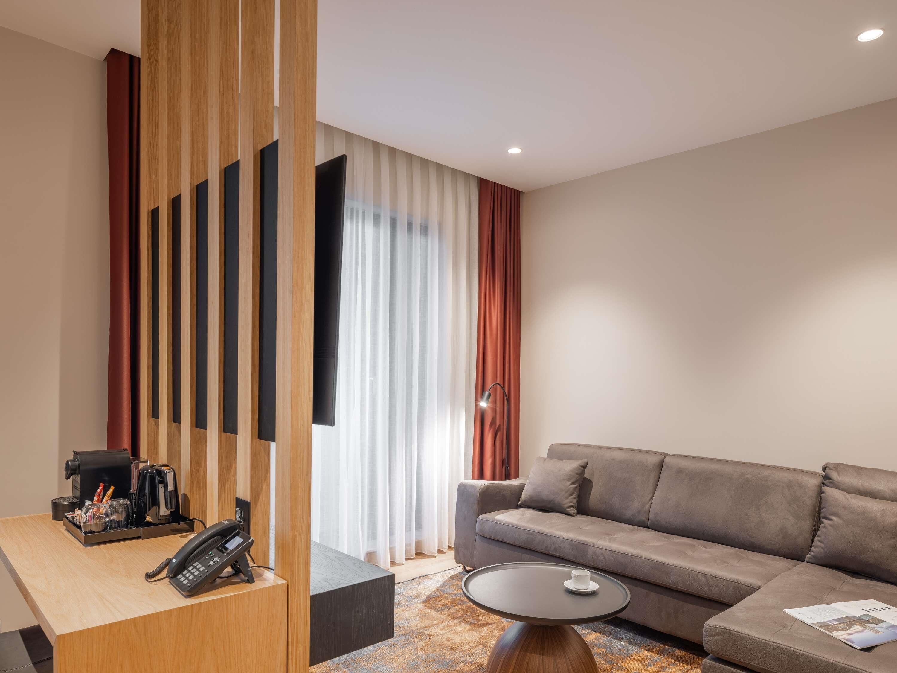 A modern hotel room with a wooden desk, a sofa, a small round table, and a large window with curtains.