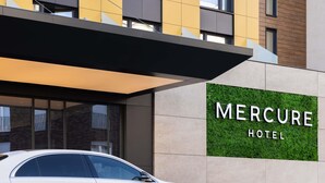 Ein weißes Auto vor dem Mercure Hotel, das ein modernes Äußeres und eine Schilder mit dem Hotelnamen hat.