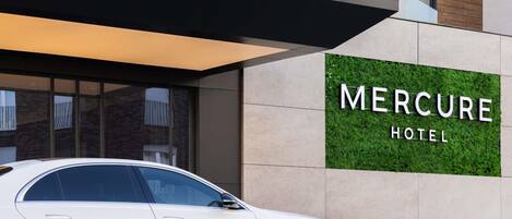 En hvid bil parkeret foran Mercure Hotel, med en moderne facade og et skilt med hotellets navn.