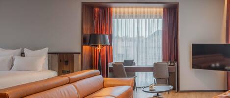 A hotel room with a bed, a leather sofa, a desk with a chair, a TV, and a large window with curtains.
