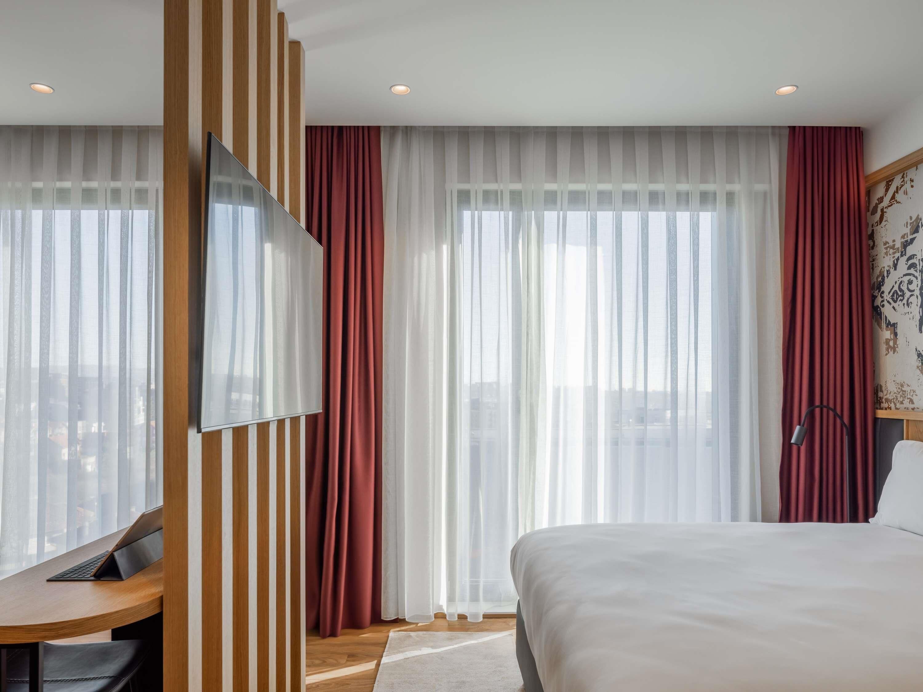 Une chambre d’hôtel avec un lit, un bureau avec un ordinateur portable, une télévision et de grandes fenêtres avec des rideaux légers.
