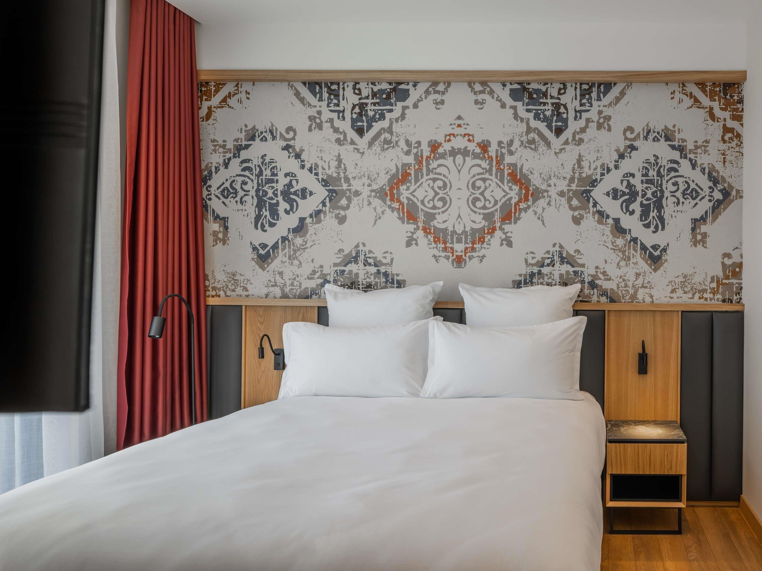 A neatly made bed with white linens, a patterned wall behind it, and a wooden nightstand with a lamp.