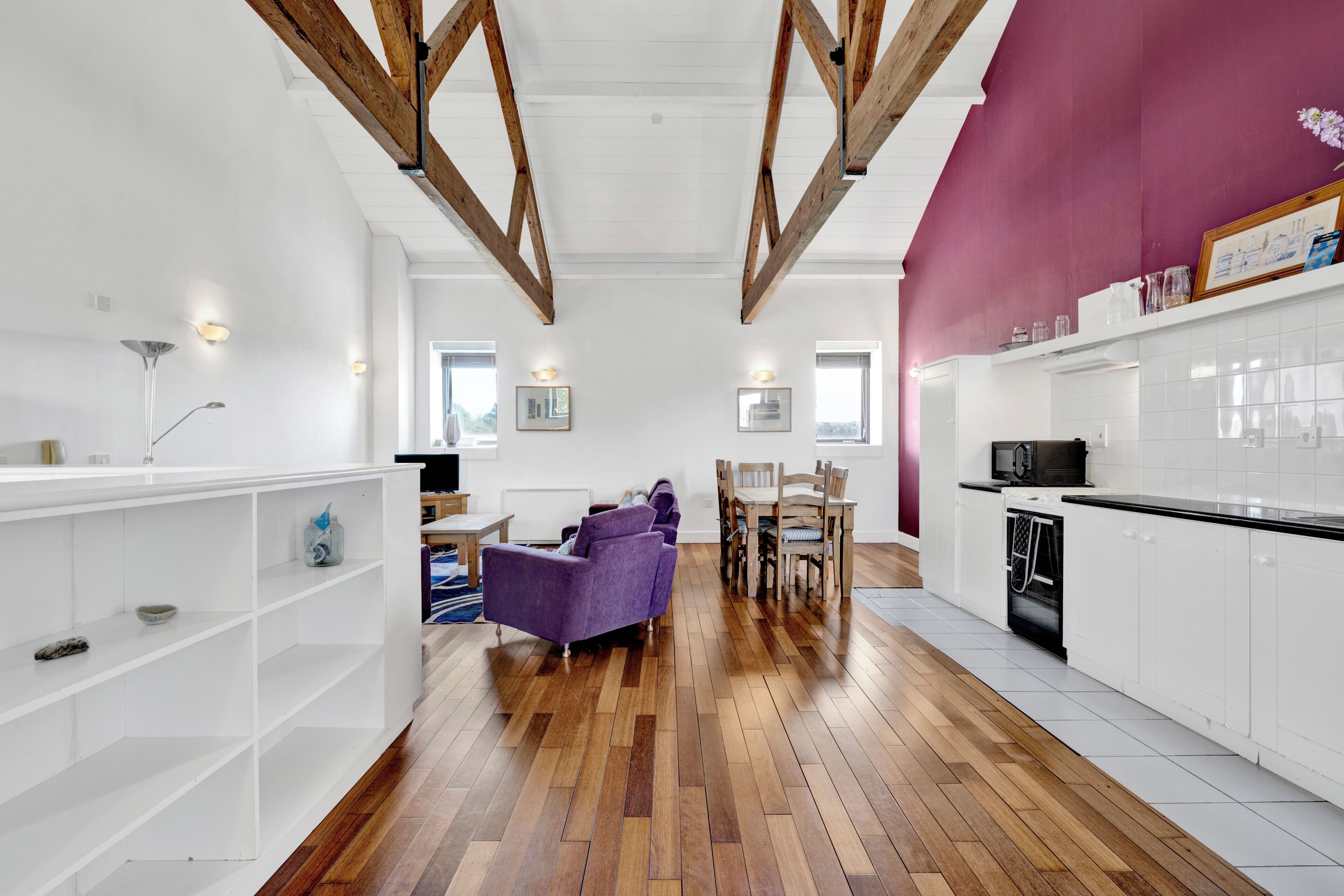 A modern kitchen with white cabinets, a dining area with a purple sofa, and wooden beams on the ceiling.