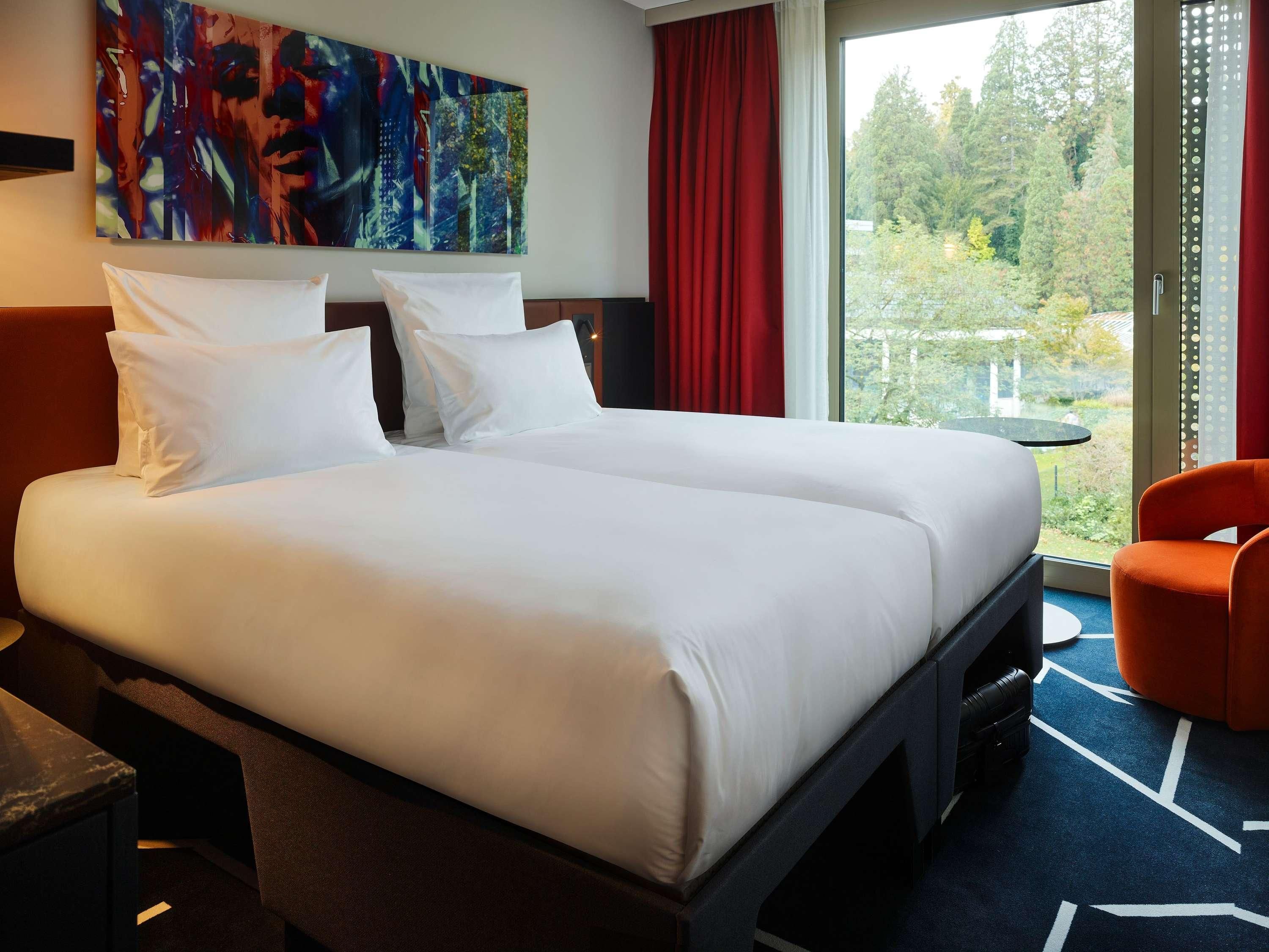 Ein Doppelbett mit weißen Leinen, ein roter Vorhang, ein großes abstraktes Gemälde und Blick auf Bäume durch ein Fenster.