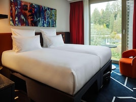 Uma cama de casal com lençóis brancos, uma cortina vermelha, uma pintura abstrata grande e vista para árvores através de uma janela.