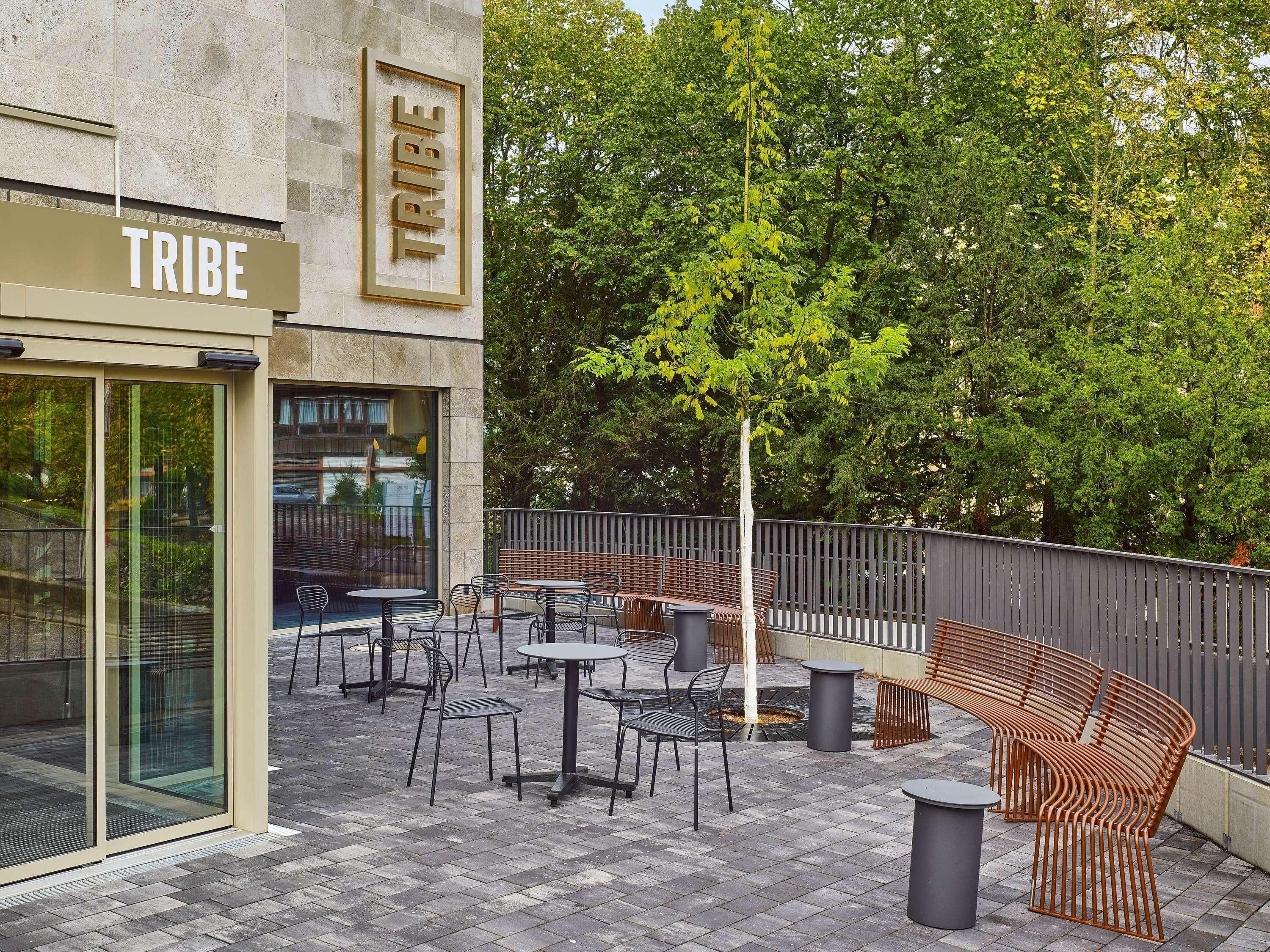 An outdoor seating area with tables and chairs, a sign reading 'TRIBE', and a tree in the background.