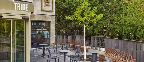 An outdoor seating area with tables and chairs, a sign reading 'TRIBE', and a tree in the background.