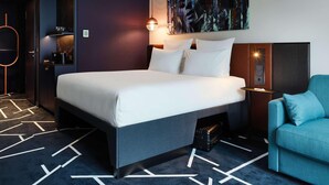 Une chambre d’hôtel moderne avec un grand lit, un fauteuil bleu, un tableau au mur et un tapis à motifs géométriques.