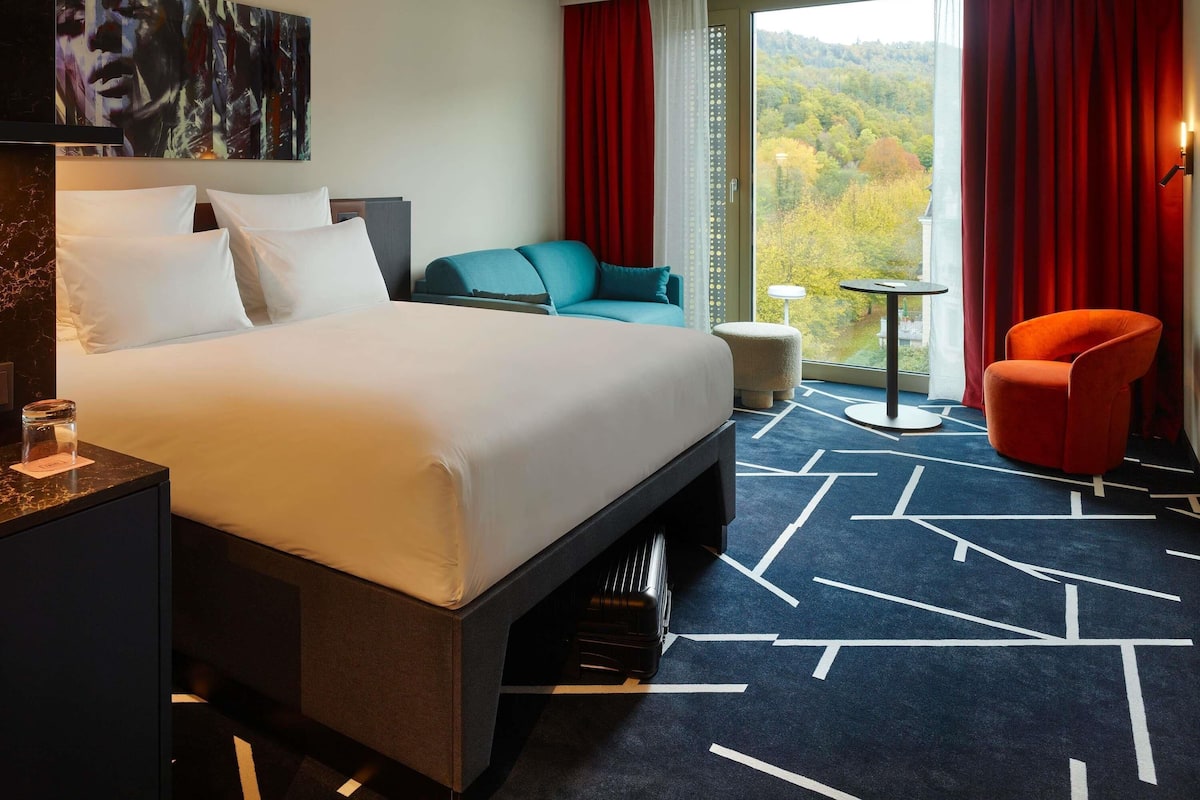 A hotel room with a large bed, a desk, a chair, a small table, and a view of a forest through the window.