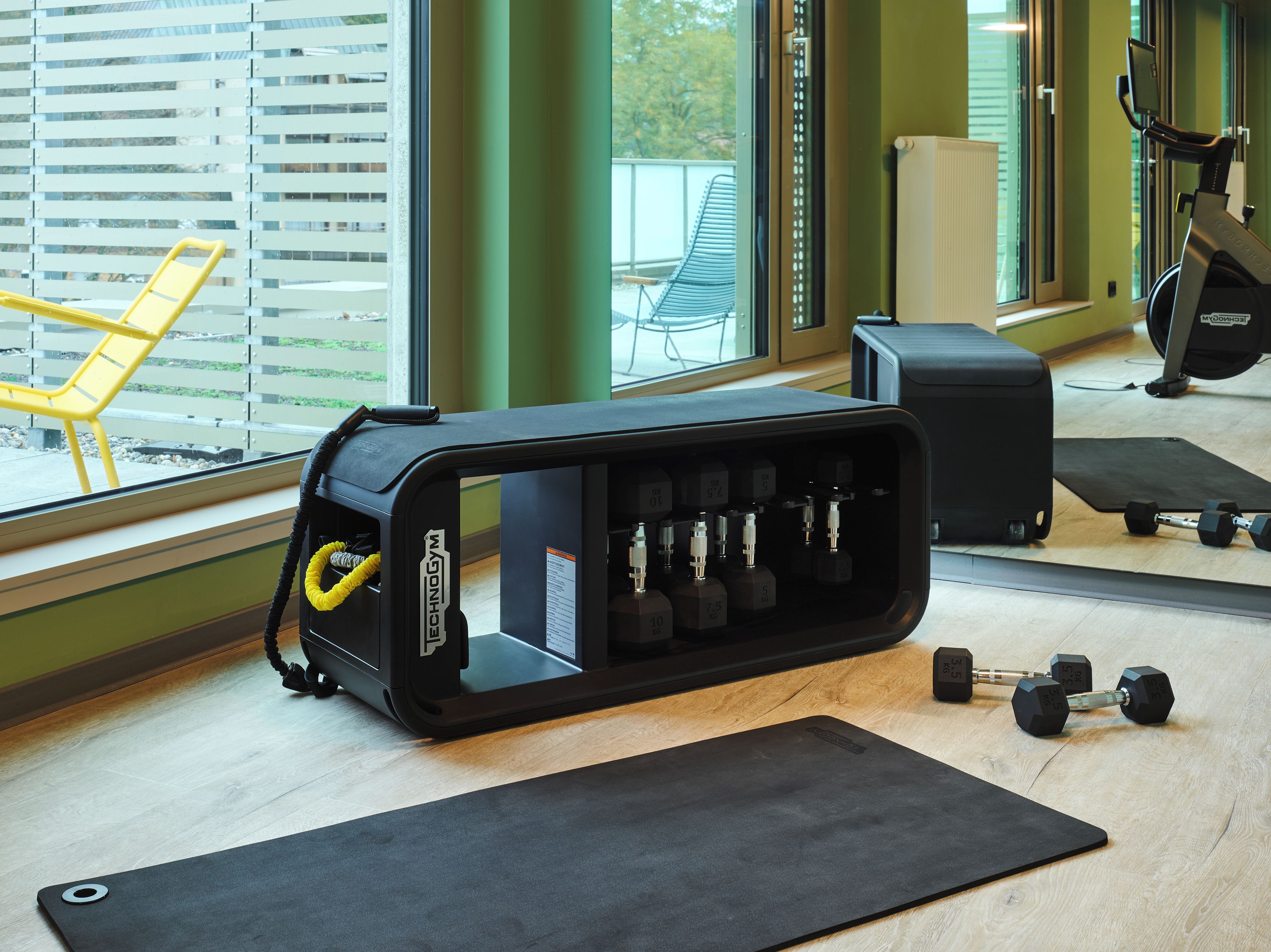 Ett gym med träningsutrustning, en yogamatta och ett fönster med persienner.