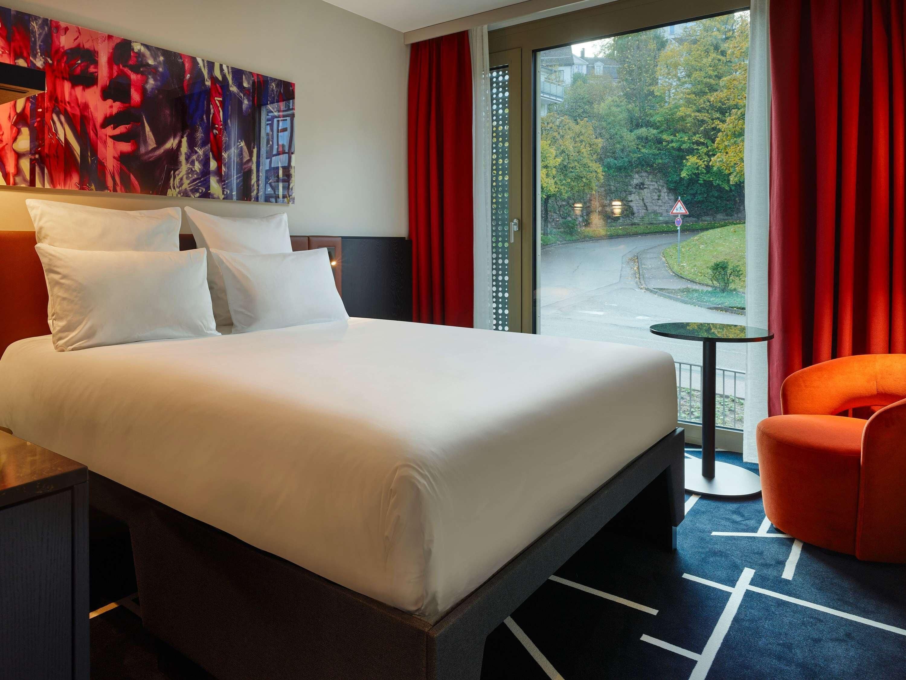 A hotel room with a large bed, a small table, a chair, and a view of a street and trees.