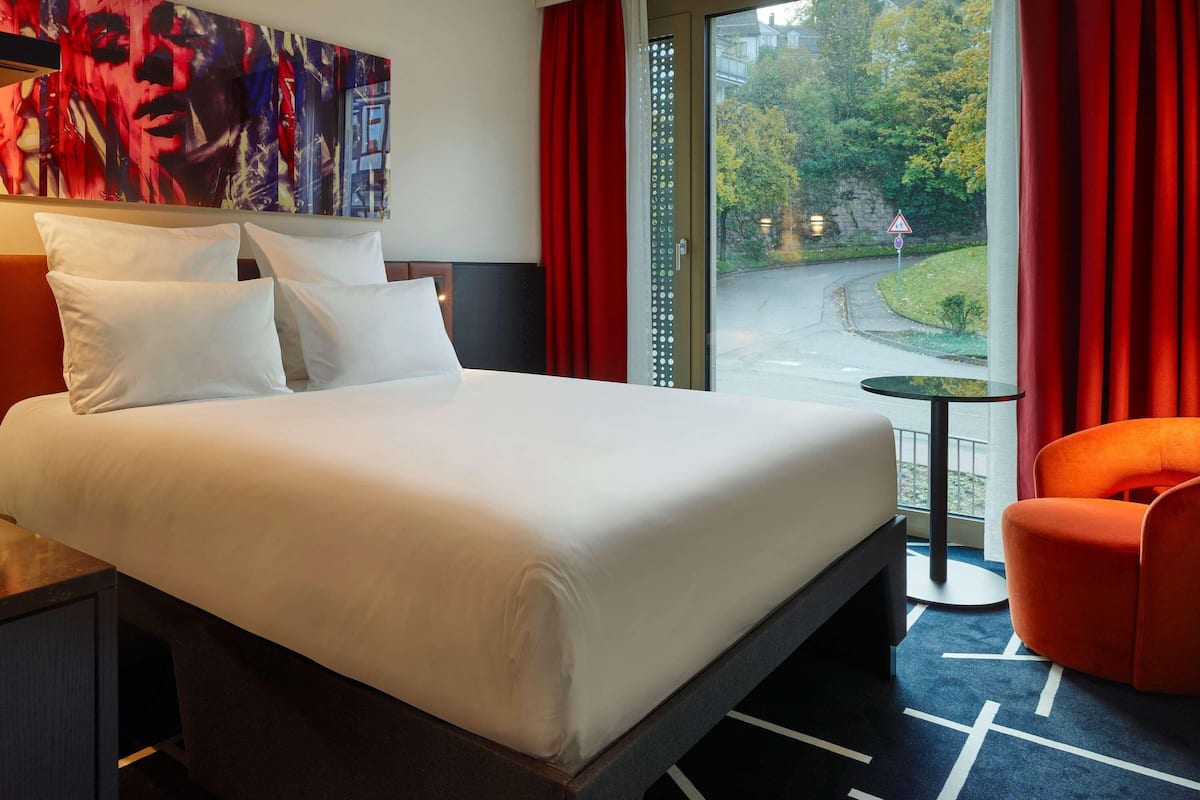A hotel room with a large bed, a small table, a chair, and a view of a street and trees.