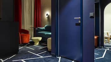 Habitación de hotel con alfombra azul oscuro, sofá color verde azulado, una silla roja, una pequeña mesa redonda y una puerta con manija.
