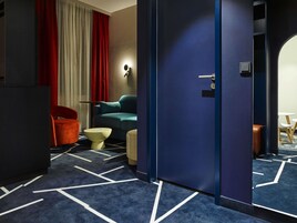 A hotel room with a dark blue carpet, a teal sofa, a red chair, a small round table, and a door with a handle.