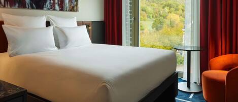 Ein Hotelzimmer mit einem großen Bett, Blick auf einen Wald, einem roten Vorhang und einem kleinen Tisch.