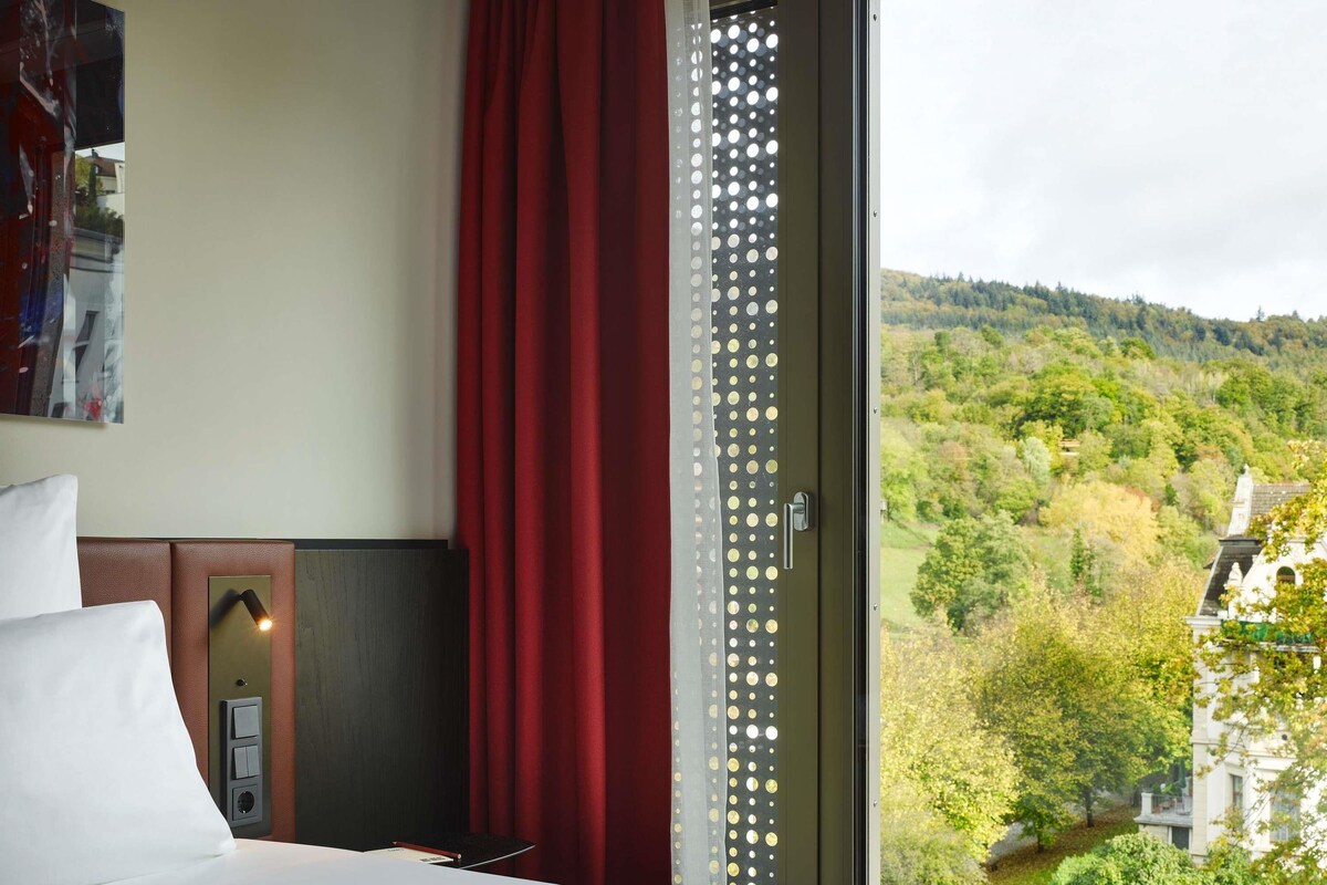A hotel room with a bed, a red curtain, a window with a view of a green landscape, and a door with a circular pattern.