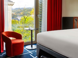 Quarto de hotel com uma cama, uma cadeira vermelha, uma mesa pequena e vista para uma paisagem verde.