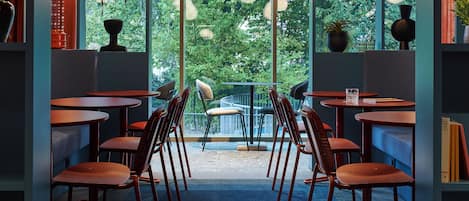 A restaurant with multiple tables and chairs, large windows with a view of trees, and hanging spherical light fixtures.
