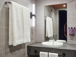 Um banheiro moderno com duas toalhas brancas penduradas em porta-toalhas, um lavatório redondo, um espelho e um luminário fixado na parede.