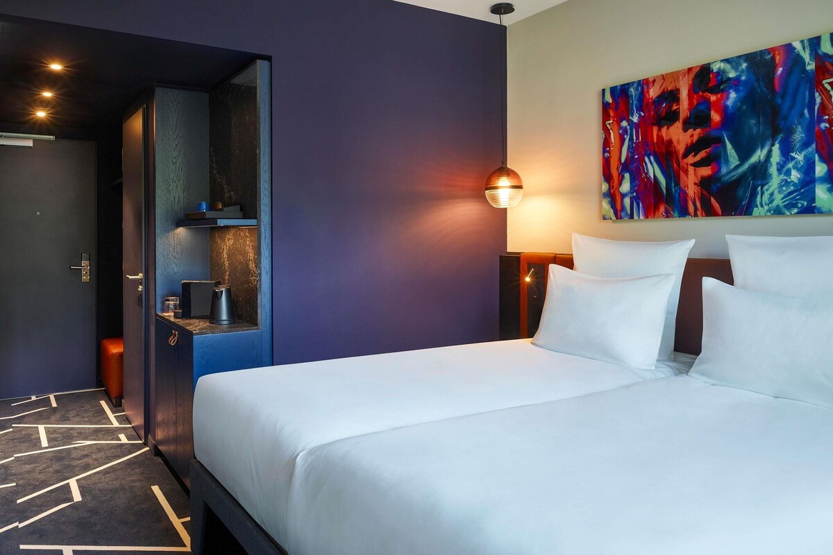 A hotel room with a bed, a painting, a nightstand, and a door leading to a bathroom.