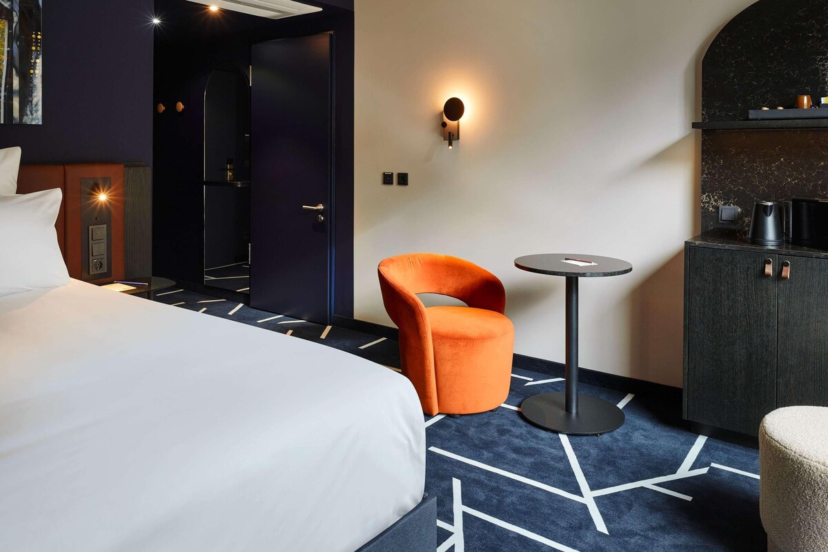 A hotel room with a bed, an orange chair, a small round table, a dark cabinet, and a wall-mounted light.