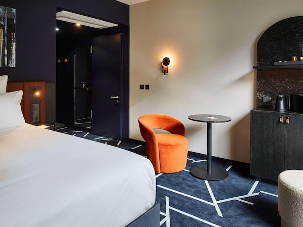 A hotel room with a bed, an orange chair, a small round table, a dark cabinet, and a wall-mounted light. - Tribe Baden Baden (Baden-Baden)