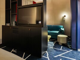 Habitación de hotel moderna con un televisor de pantalla plana grande, un sofá color turquesa, una pequeña mesa redonda y una alfombra estampada.
