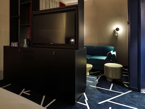 A modern hotel room with a flat-screen TV, a sofa, a small round ottoman, and a patterned carpet. - Tribe Baden Baden (Baden-Baden)