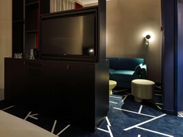 Quarto de hotel moderno com televisão de tela plana, sofá, um banquinho redondo pequeno e carpete estampado.