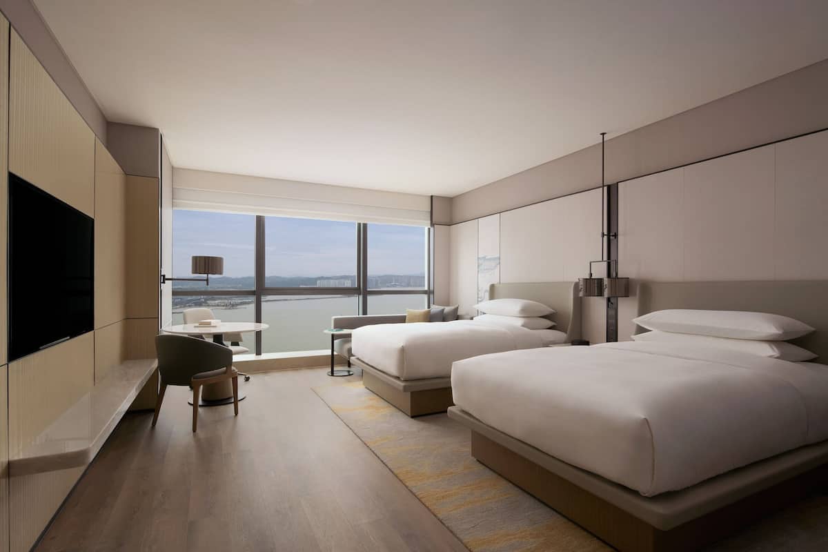 A modern hotel room with two beds, a flat-screen TV, a desk, and a view of the city.