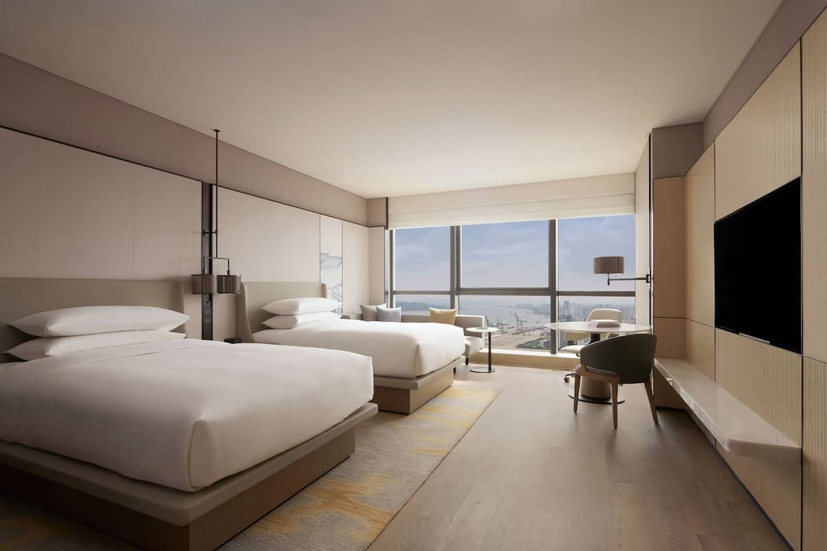 A modern hotel room with two large beds, a small seating area, and a large window offering a city view.