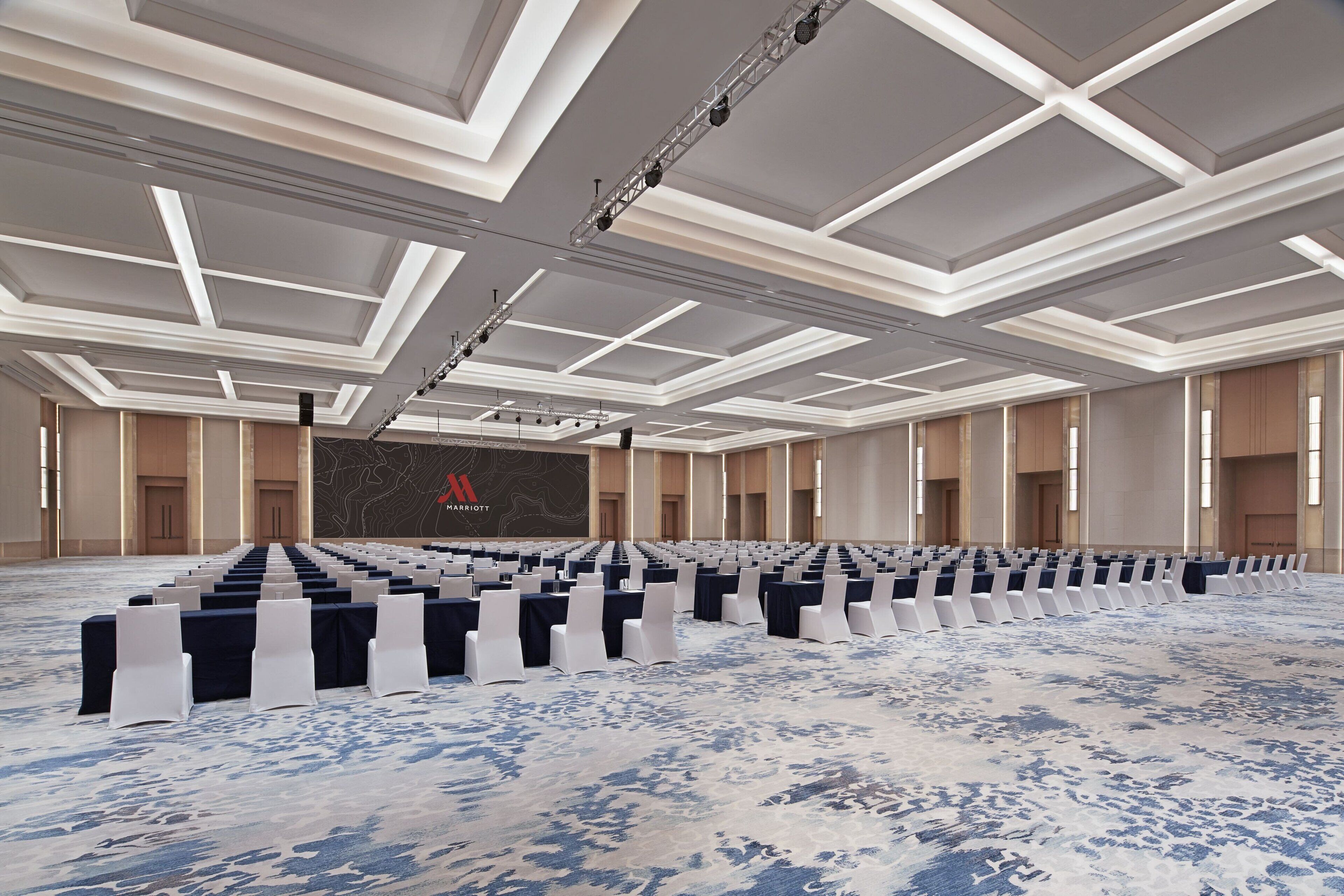 Un amplio salón de conferencias con filas de mesas y sillas, un diseño de techo moderno y una pantalla grande al frente.