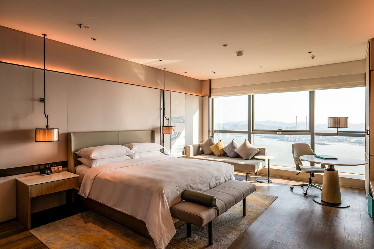 A modern hotel room with a large bed, a desk, a chair, and a view of the city.