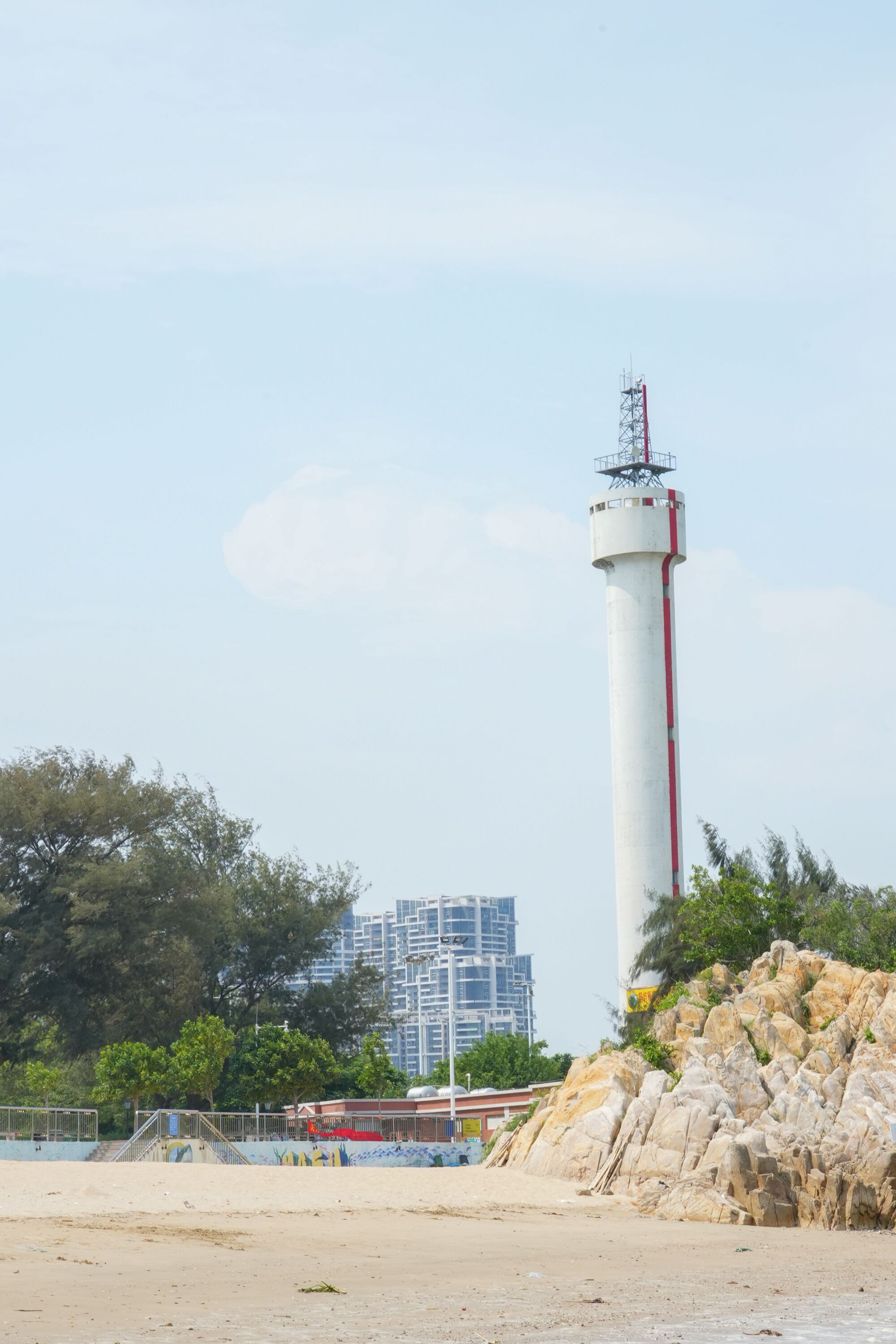A lighthouse with a white and red structure, situated on a sandy beach with rocky outcrops and trees in the background.